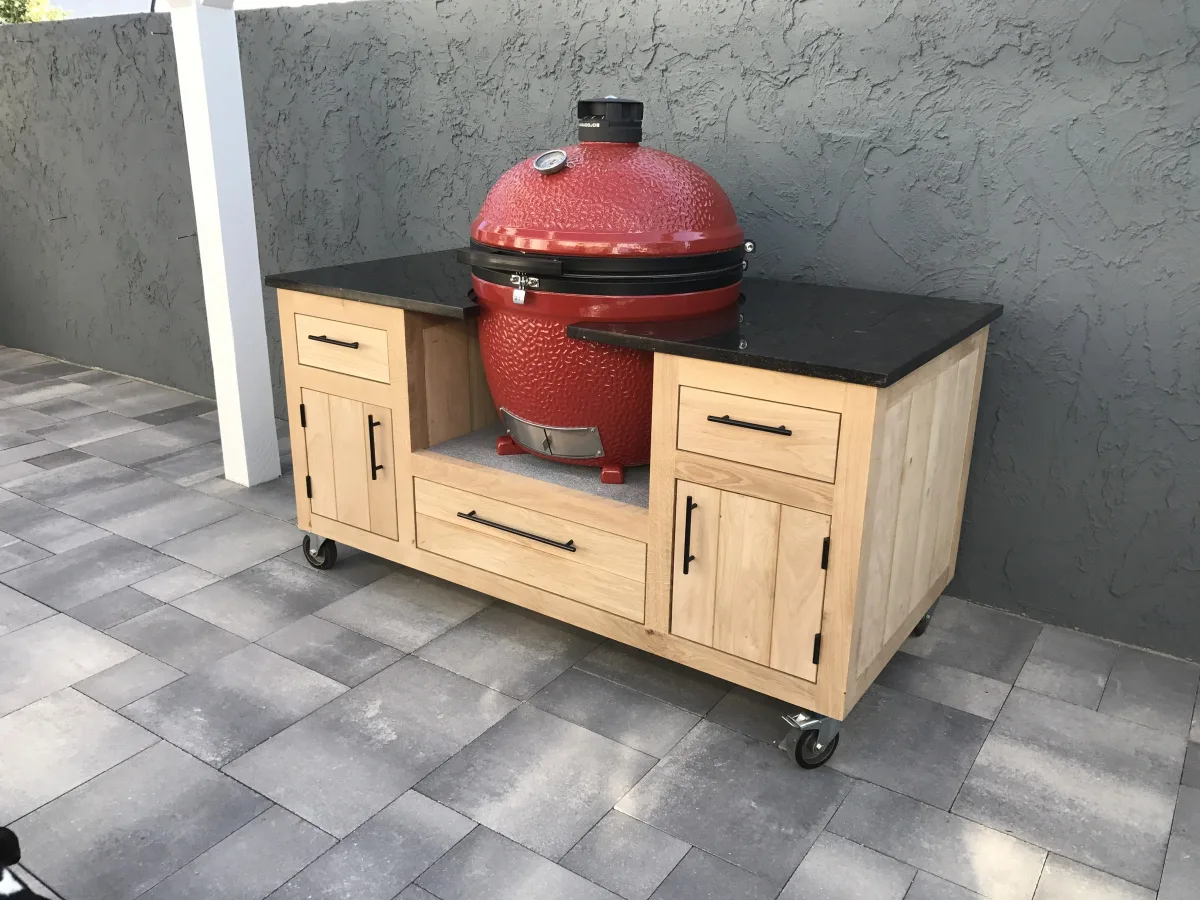 Kamado Buitenkeuken Maatwerk buitenkeuken, Verrijdbare eiken buitenkeuken met zwart natuurstenen werkblad en rode Kamado Joe, driekwart van linksvoor op een terras