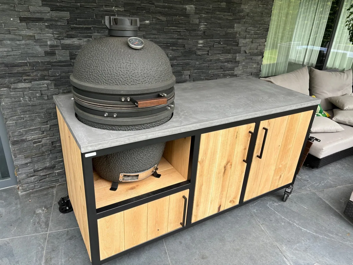 MB Industrial Living Basic, Buitenkeuken Basic met zwart stalen frame, eiken fronten en grijs Mortex werkblad, kamado links en twee houten deuren, driekwart van linksvoor op een overdekt terras