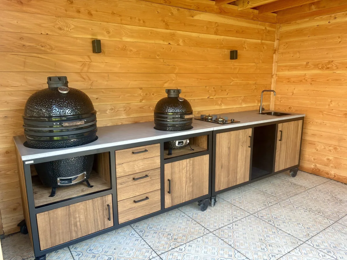 MB Industrial Living Maatwerk buitenkeuken, Maatwerk buitenkeuken met zwart stalen frame, eiken fronten en grijs werkblad, twee kamados, kookplaat en spoelbak, in een houten veranda