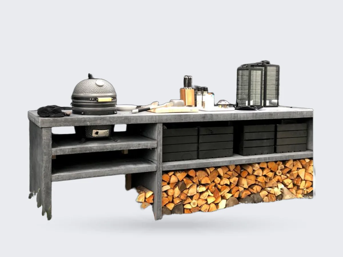 Pronck Buitenkeuken Beton, Lange betonnen buitenkeuken met rokende kamado, open vakken, zwarte kratten en houtopslag voor een zwarte bakstenen muur