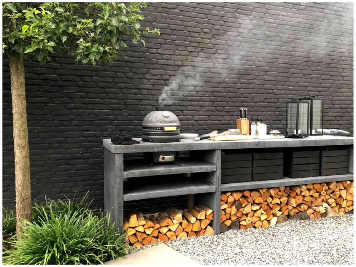 Pronck Buitenkeuken Beton, Lange betonnen buitenkeuken met rokende kamado, open vakken, zwarte kratten en houtopslag voor een zwarte bakstenen muur