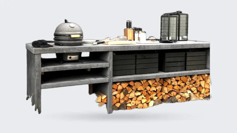 Pronck Buitenkeuken Beton, Lange betonnen buitenkeuken met rokende kamado, open vakken, zwarte kratten en houtopslag voor een zwarte bakstenen muur