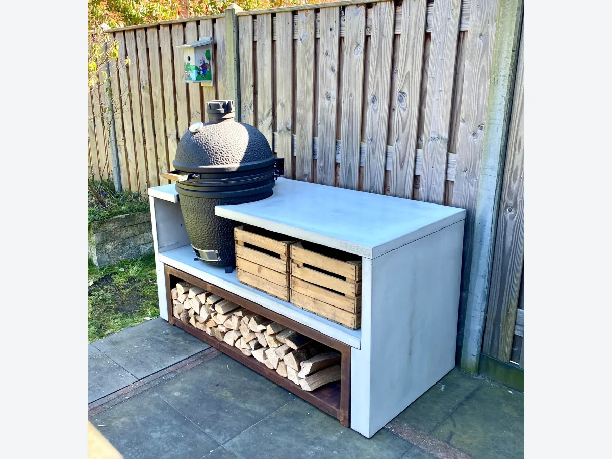 STEGG STEGG buitenkeuken, Grijze STEGG buitenkeuken met zwarte kamado, kratten en cortenstalen houtopslag voor een houten schutting