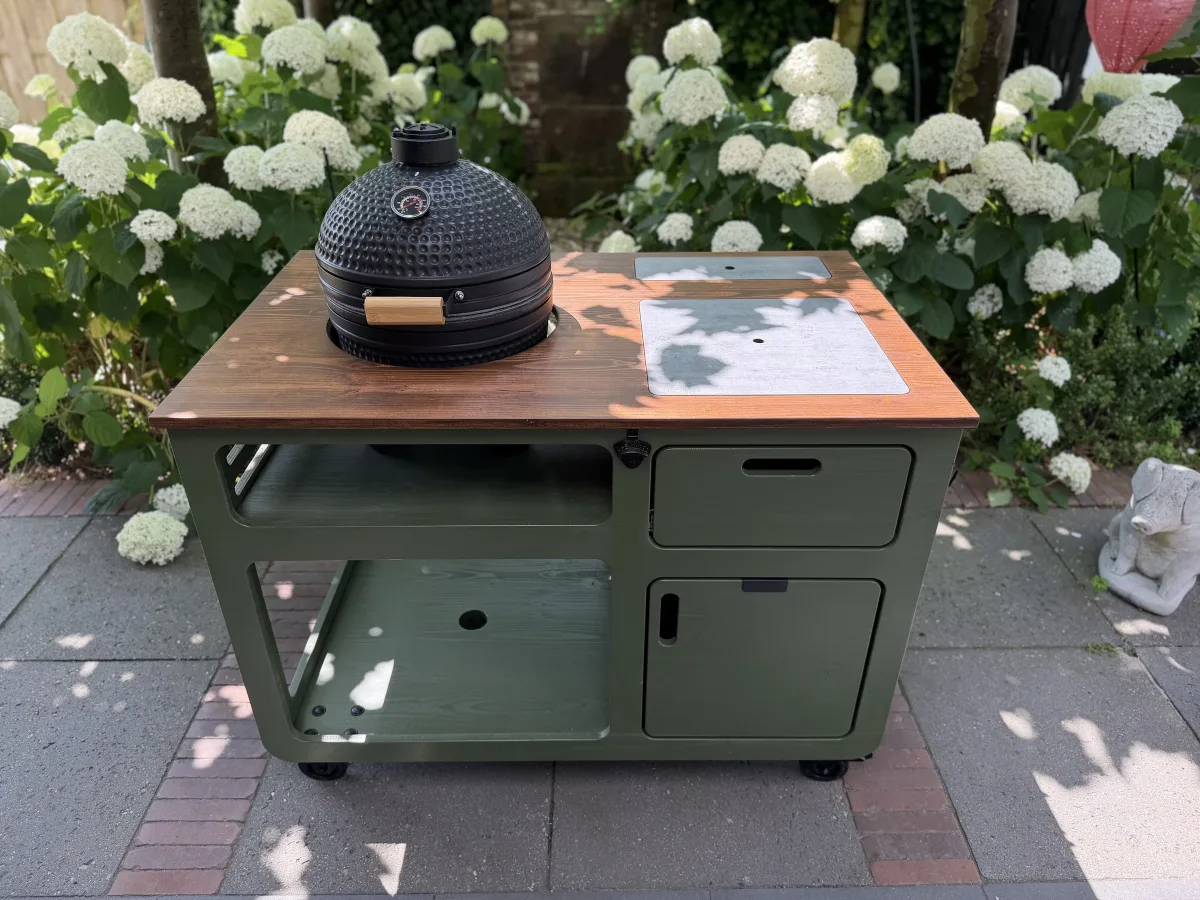 Studio RUUB Olyv buitenkeuken, Olyv buitenkeuken van olijfgroen douglas met zwarte kamado, hittebestendige inlay, lade en deur, driekwart van linksvoor op een terras tussen hortensia's