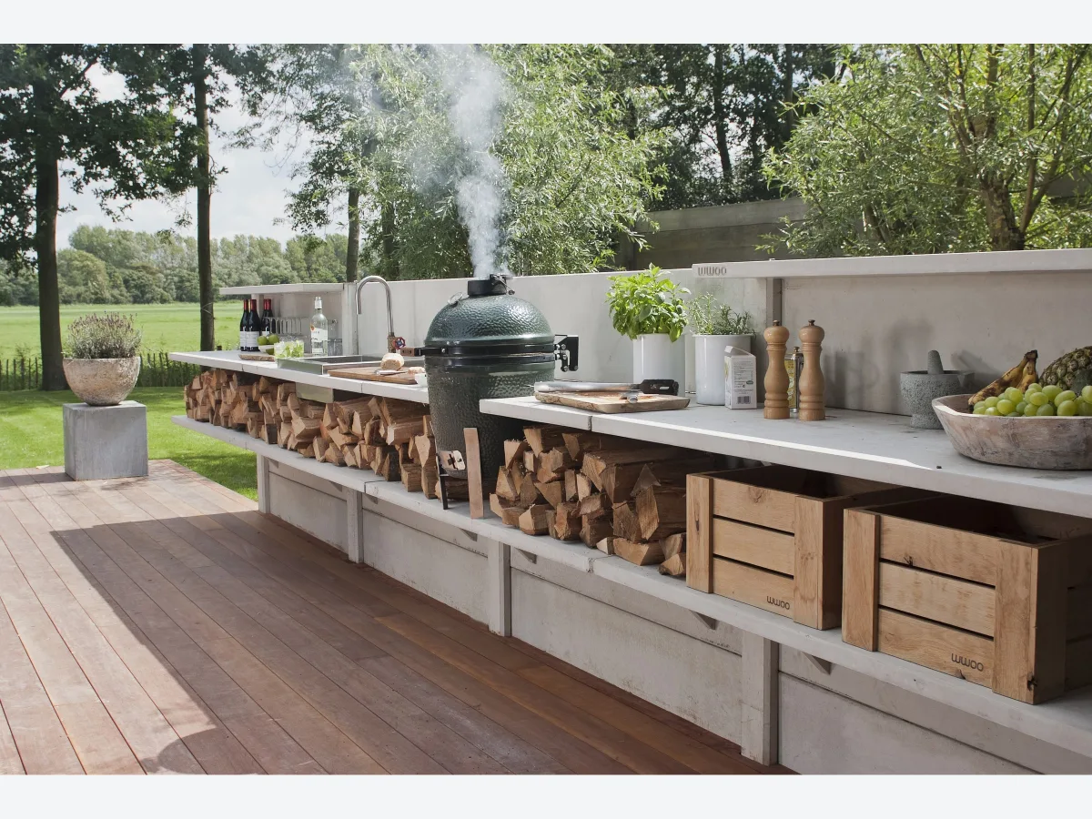 WWOO Beton, WWOO betonnen buitenkeuken in grijs op een houten vlonder met rokende Big Green Egg, haardhout en houten kratten