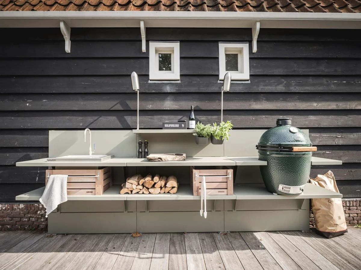 WWOO Staal, WWOO stalen buitenkeuken in groen met spoelbak, haardhout, houten kratten en Big Green Egg, tegen een zwarte houten schuur